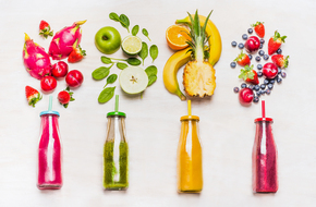 4 leżące butelki soków w różnych kolorach, a nad nimi owoce, pitahaja, jabłko, banan, ananas, truskawki, borówki