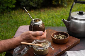 Naczynie pełne naparu yerba mate trzymane w męskiej dłoni, najwyraźniej w czasie pikniku, w tle stalowy czajnik, jeszcze dalej widoczna zielona trawa.