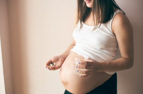 Kobieta w ciąży trzyma w jednej dłoni paracetamol a w drugiej szklankę wody.