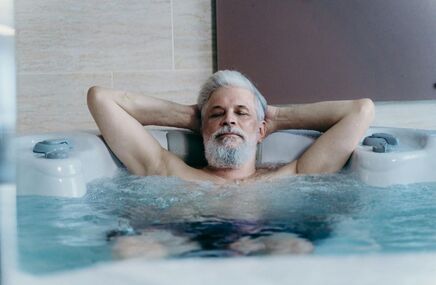 Starszy mężczyzna z siwymi włosami i brodą relaksuje się w jacuzzi z zamkniętymi oczami. Ręce ma założone za głowę, woda bulgocze wokół niego. 
