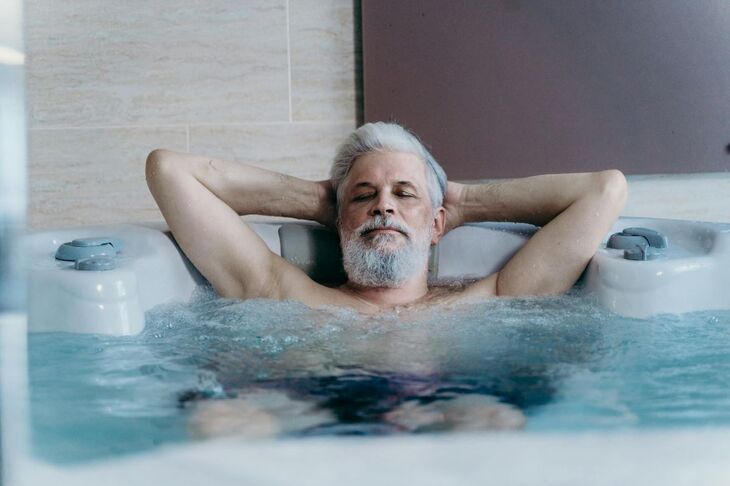 Starszy mężczyzna z siwymi włosami i brodą relaksuje się w jacuzzi z zamkniętymi oczami. Ręce ma założone za głowę, woda bulgocze wokół niego. 
