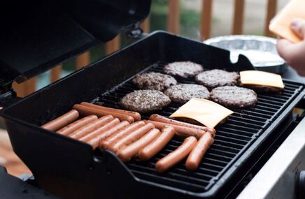 Grill z hamburgerami, kiełbaskami i plastrami sera grillującymi się na ruszcie. Ręka trzyma łopatkę do grillowania. Letni barbecue na zewnątrz.