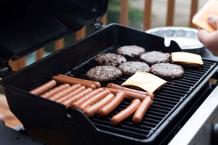 Grill z hamburgerami, kiełbaskami i plastrami sera grillującymi się na ruszcie. Ręka trzyma łopatkę do grillowania. Letni barbecue na zewnątrz.